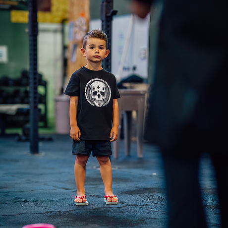 Northern Spirit presents a 4-year-old child wearing a black NS BALDER KID t-shirt with a white skull on the chest. The child is inside a sports box, listening to their coach's briefing.