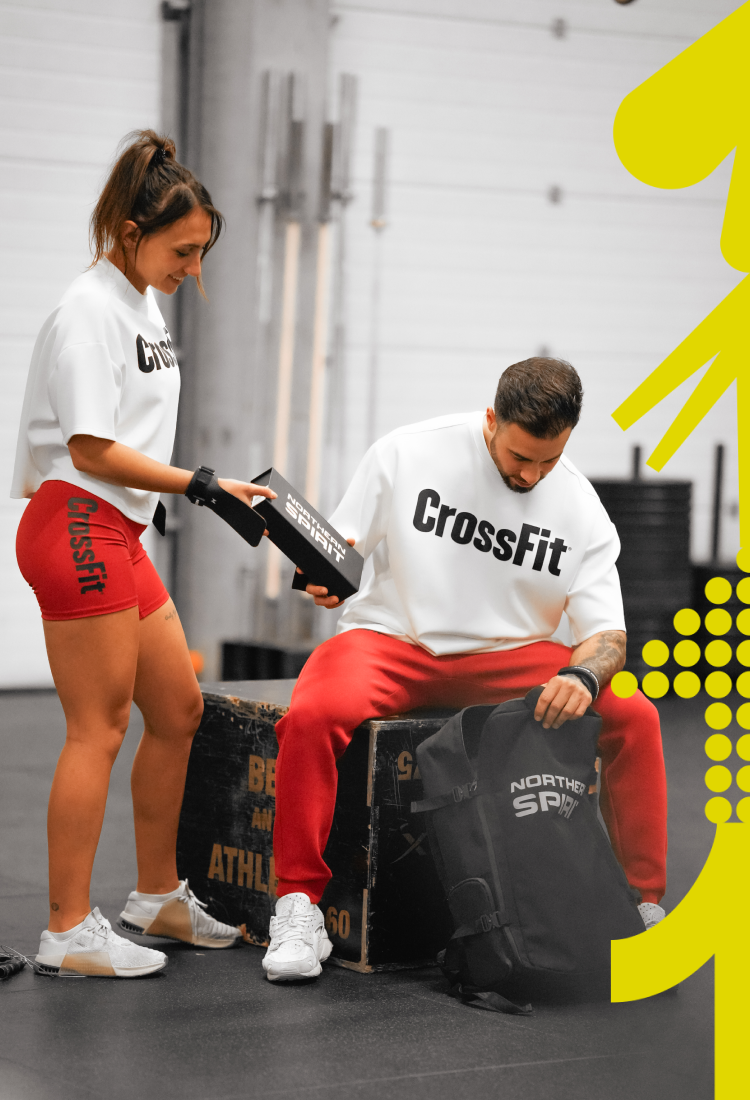 CrossFit athletes wearing white CrossFit® tops and red shorts inside a CrossFit gym, preparing their training gear with Northern Spirit backpack and accessories between workouts