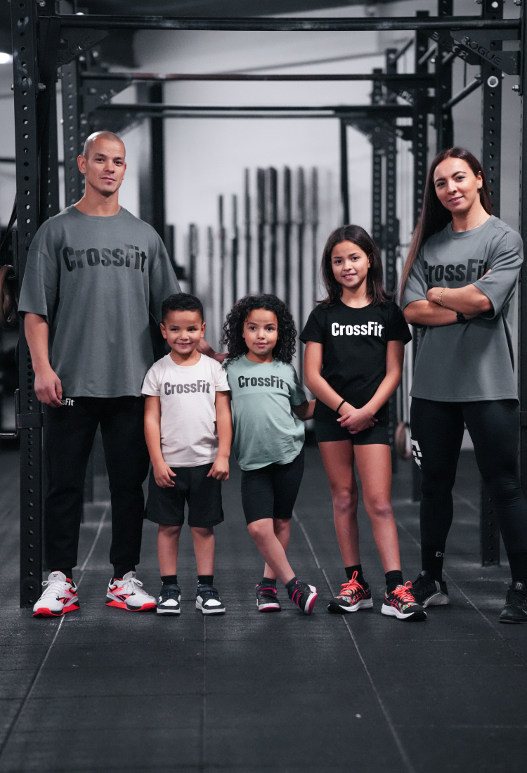 Adults and children standing together in a CrossFit gym wearing CrossFit t-shirts, showcasing a family-friendly functional fitness atmosphere.