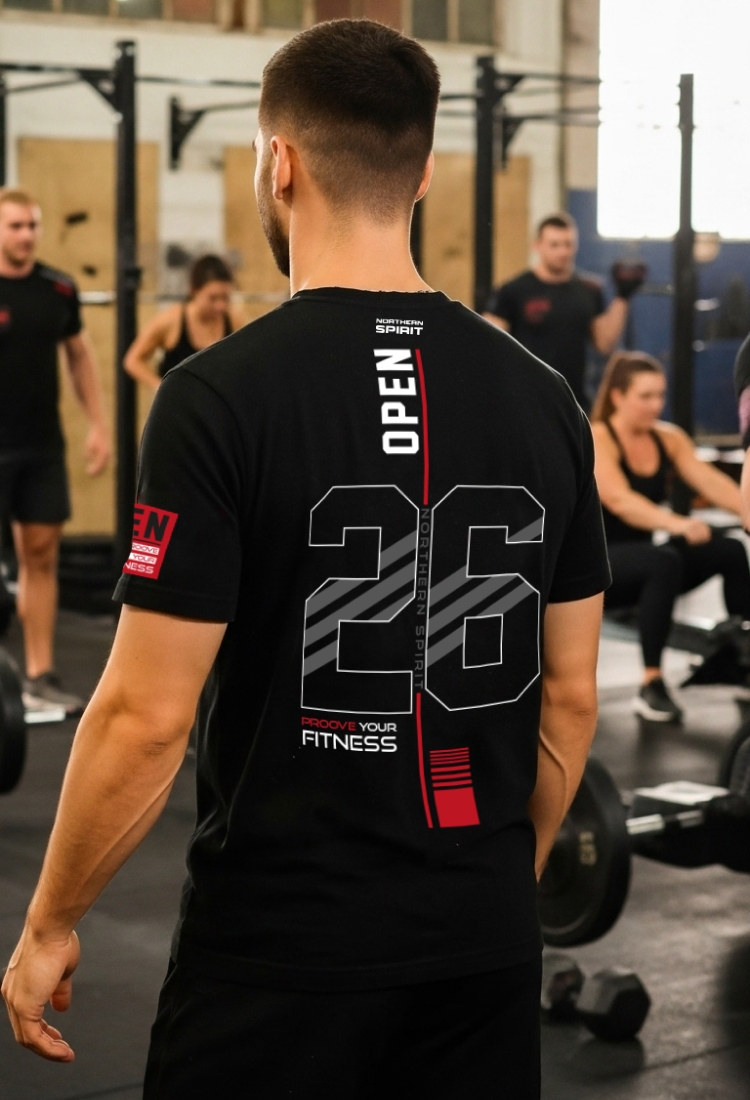 Man wearing a black Northern Spirit Open 26 CrossFit® t-shirt seen from the back in a CrossFit gym, featuring a large 26 Open graphic and “Prove Your Fitness” print during a group workout session.
