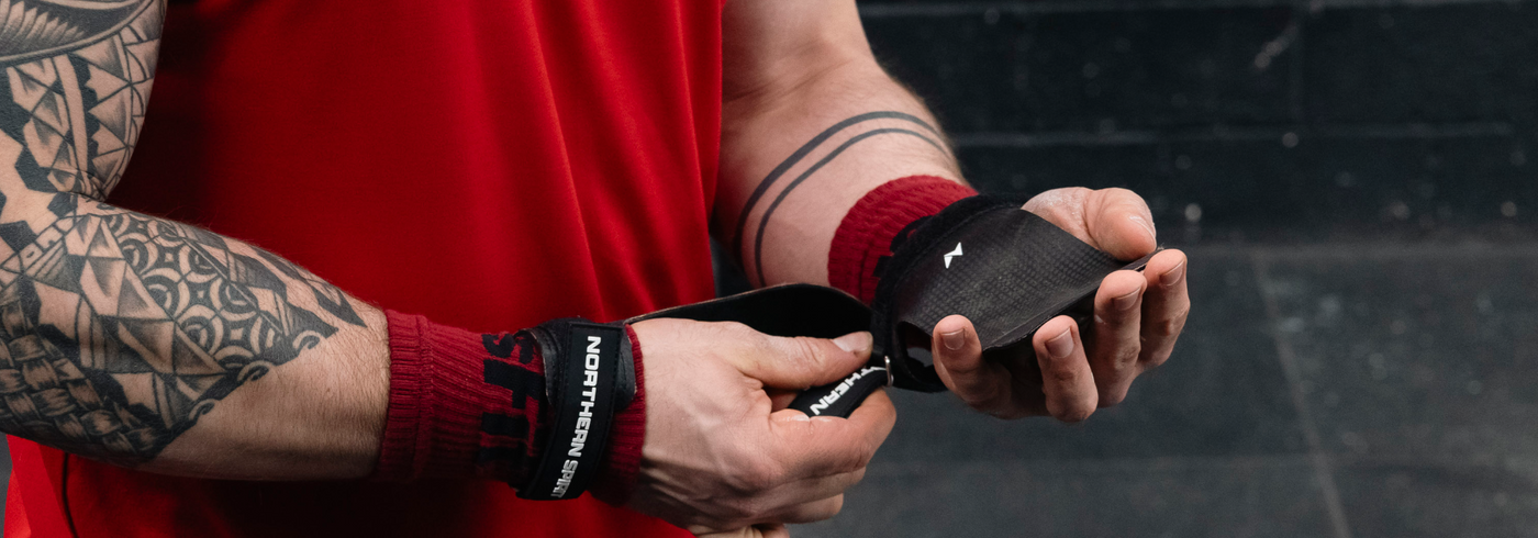 Male CrossFit® athlete wearing red shirt adjusting Northern Spirit black hand grips and wrist wraps before training in a gym.