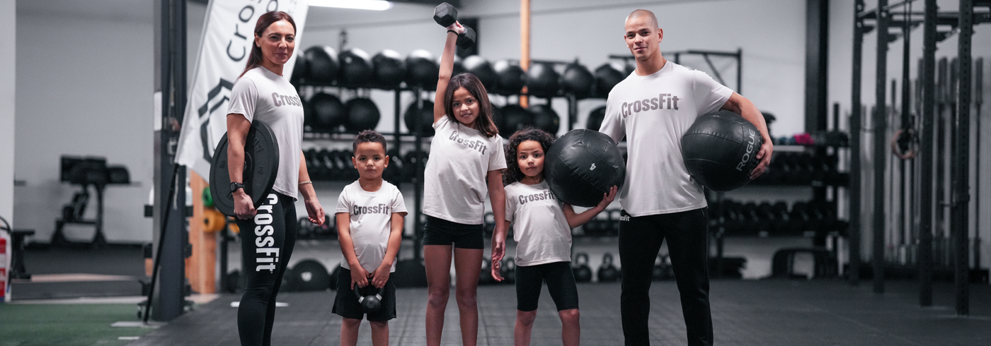 Family training together in a CrossFit gym wearing CrossFit apparel, adults and kids holding workout equipment in a functional fitness environment.