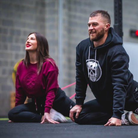 Two athletes are doing mobility exercises. They both are wearing outfits from the Colorskull collection: black joggers, purple crop hoodies, and black hoodies.