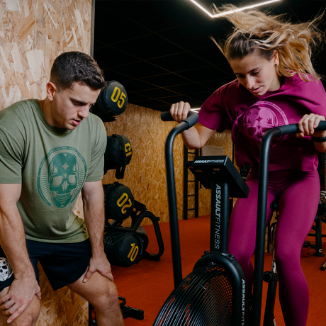 Two athletes are putting in a lot of effort, one on the assault bike, and the other cheering them on. They're dressed in the Skull collection by Northern Spirit.