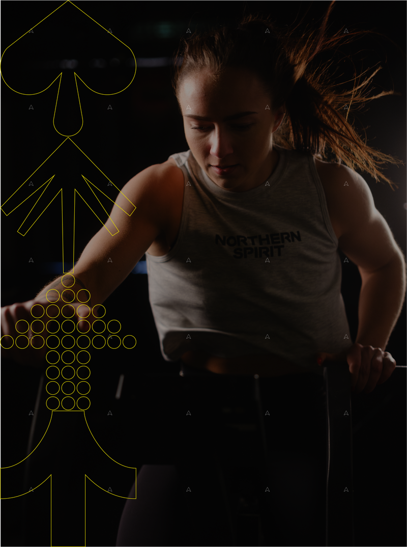 Female athlete training on an air bike wearing Northern Spirit Core Collection tank top in a dark CrossFit® gym background.