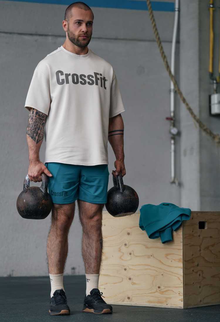 Athlete performing a farmer carry in a CrossFit gym while wearing the Northern Spirit oversized CrossFit® T-shirt in beige and teal training shorts, showcasing movement-ready apparel and functional fitness performance.