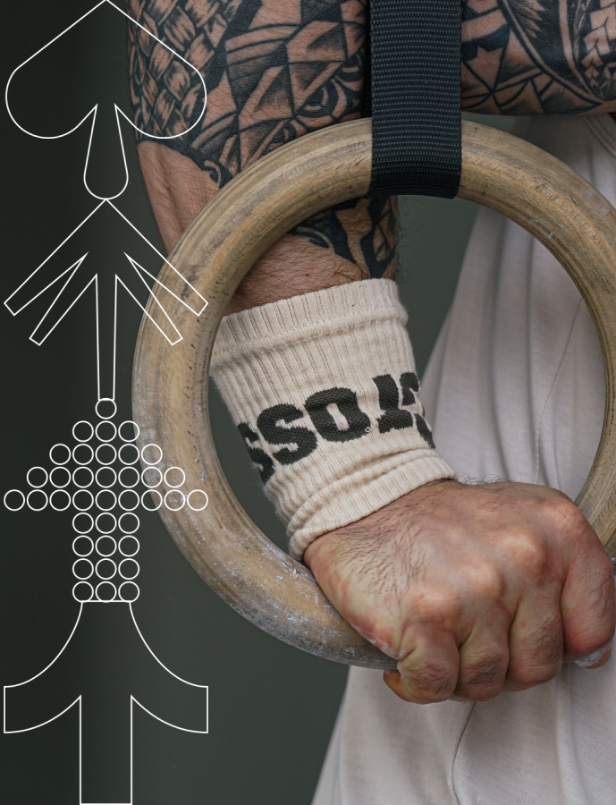 Person holding a wooden gymnastic ring with tattoos and a wristband on a dark background